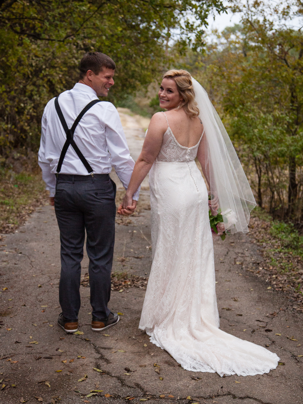 Everlasting Elopements couple looking over their shoulders back at the camera smiling on a paved path with trees at Chapel In The Oaks