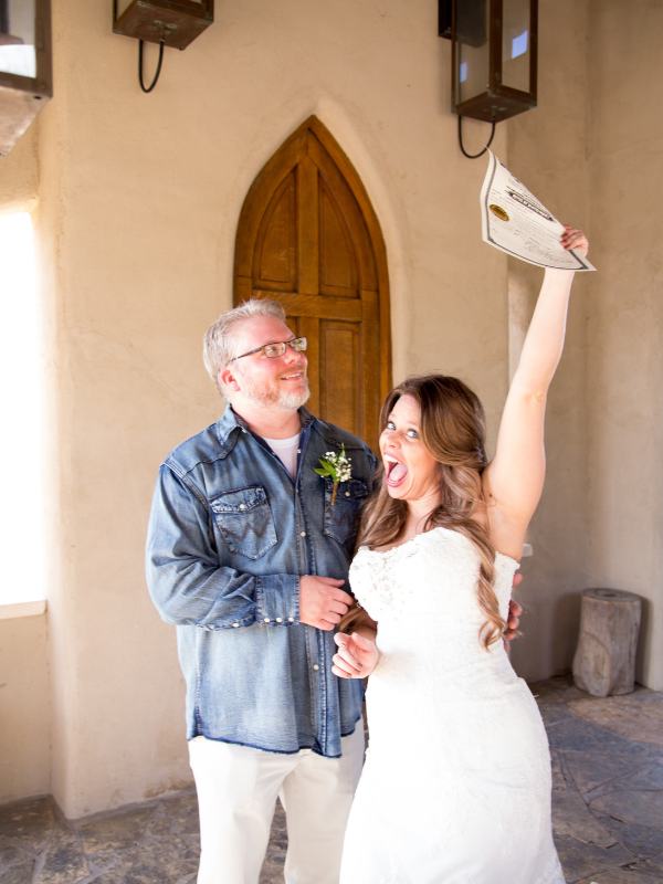 Everlasting Elopements couple smiling and the bride is holding up their marriage license in front of a door at Chapel Dulcinea 